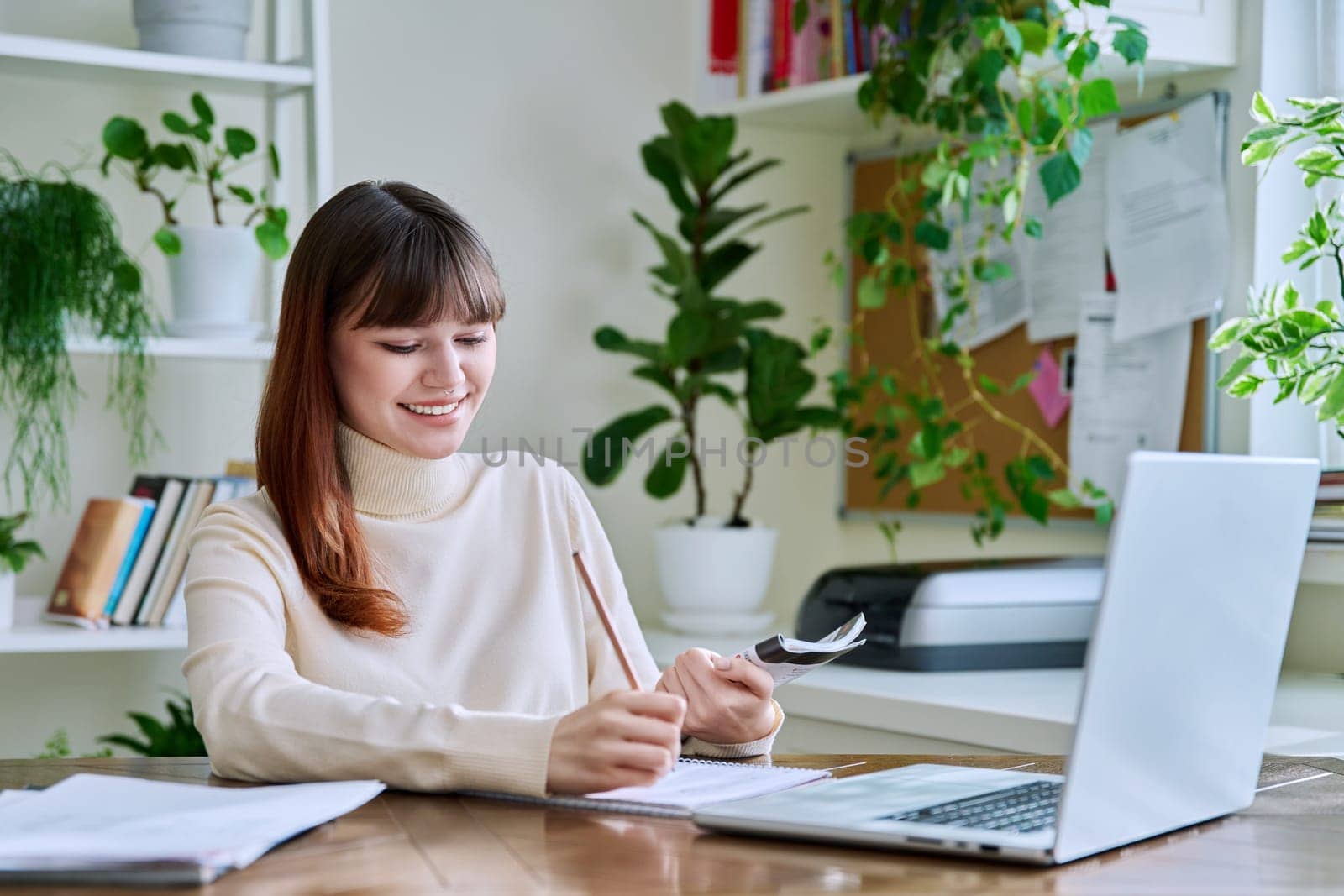 Young female college student studying at home at desk using computer laptop, writing in notebook. E-learning, education, technology, knowledge, youth concept