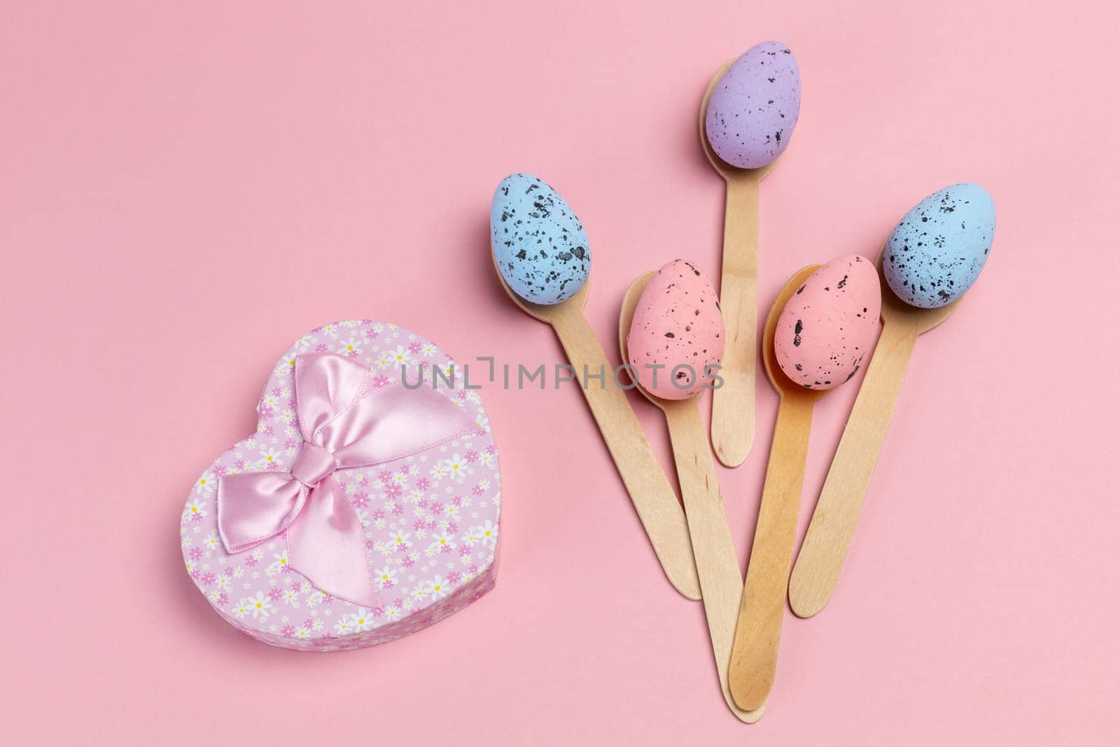 Colored Easter eggs on wooden spoons with a gift box on the pink background. Top view.