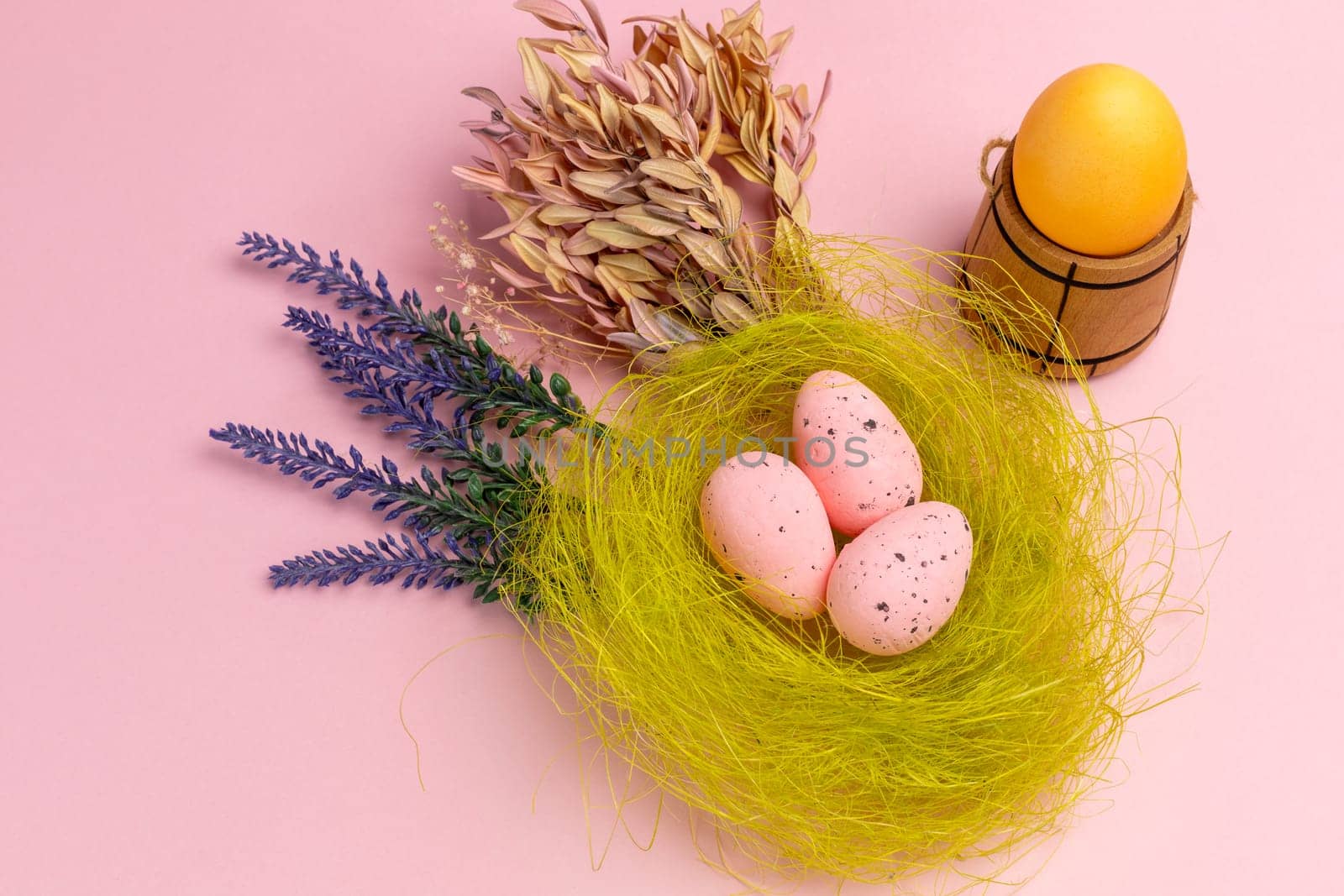 Nest with colored Easter eggs on the pink background with decor plants and wooden utensil. Top view.
