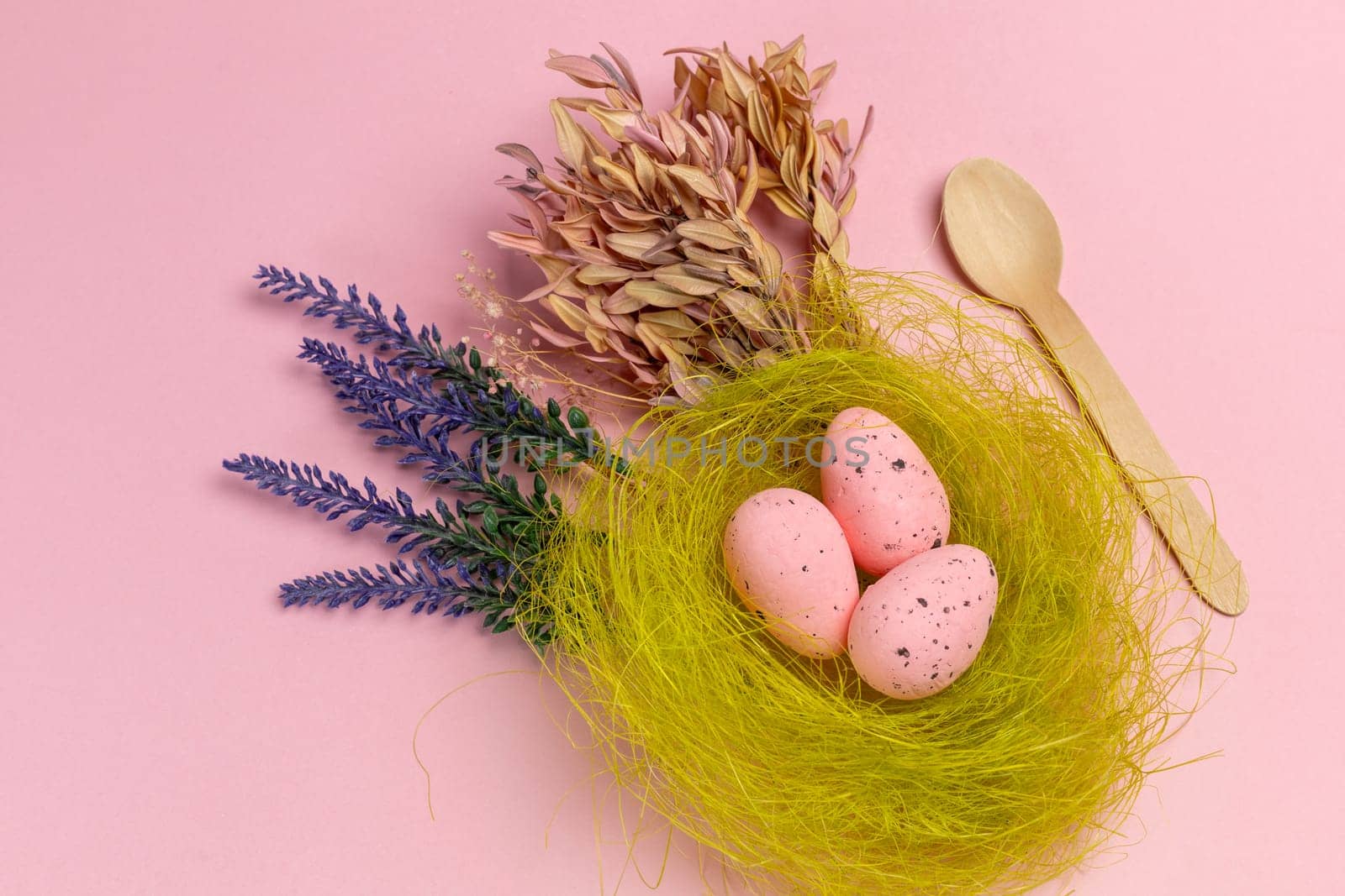 Nest with pink Easter eggs on the pink background with decor plants and a wooden spoon. Top view.