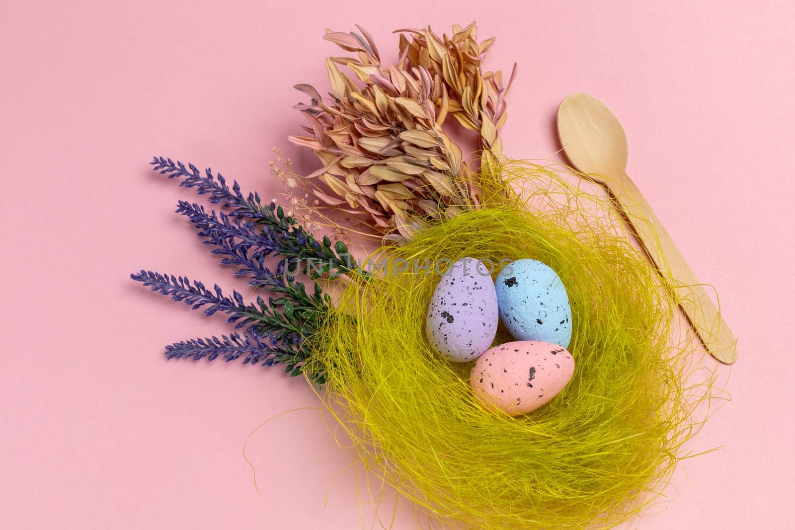 Nest with colored Easter eggs on the pink background with decor plants and a wooden spoon. Top view.