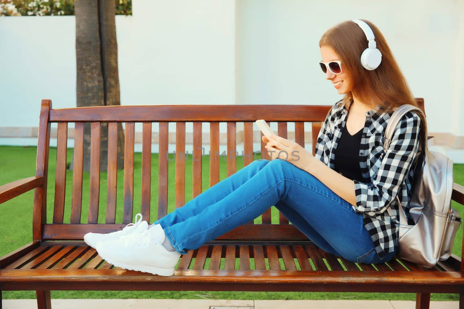 young woman listening to music in headphones with smartphone sitting on a bench in the summer park by Rohappy