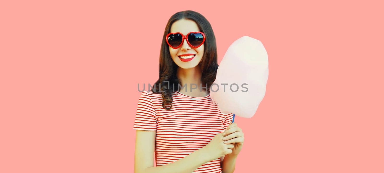 Portrait of happy smiling young woman with cotton candy wearing red heart shaped sunglasses on pink background