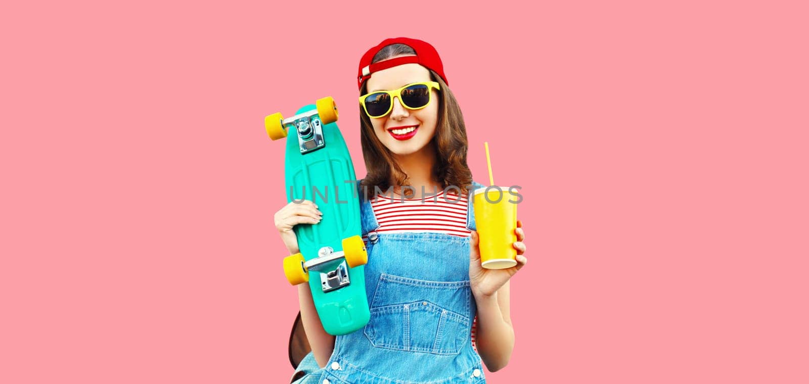 Portrait of happy smiling young woman with skateboard and cup of juice wearing red baseball cap, sunglasses on pink background