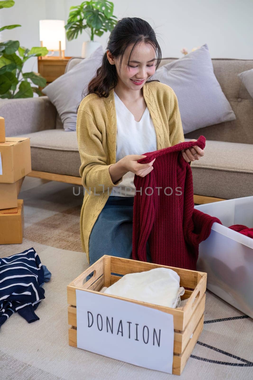 Cute Asian woman sitting next to sofa in living room sorting unwanted clothes for donation. by wichayada