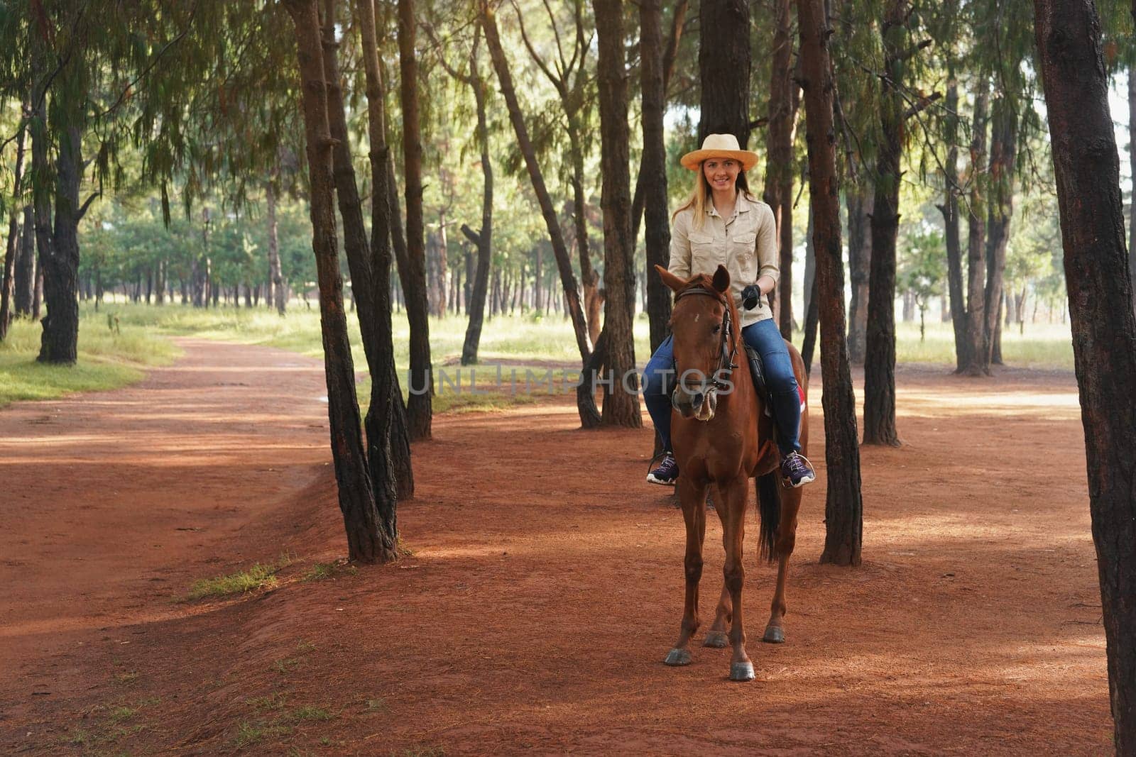 Young woman in shirt and straw hat, riding brown horse on park path, smiling, blurred trees in background
