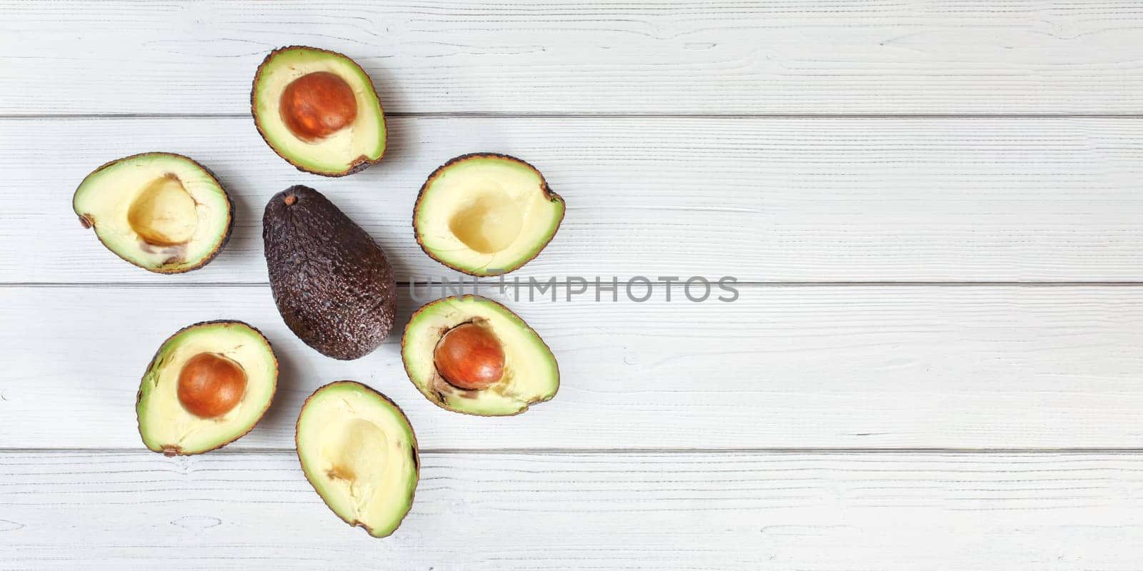 Ripe avocado halves arranged around whole dark brown pear (hass / bilse fruit variety) on white boards desk, banner with space for text right