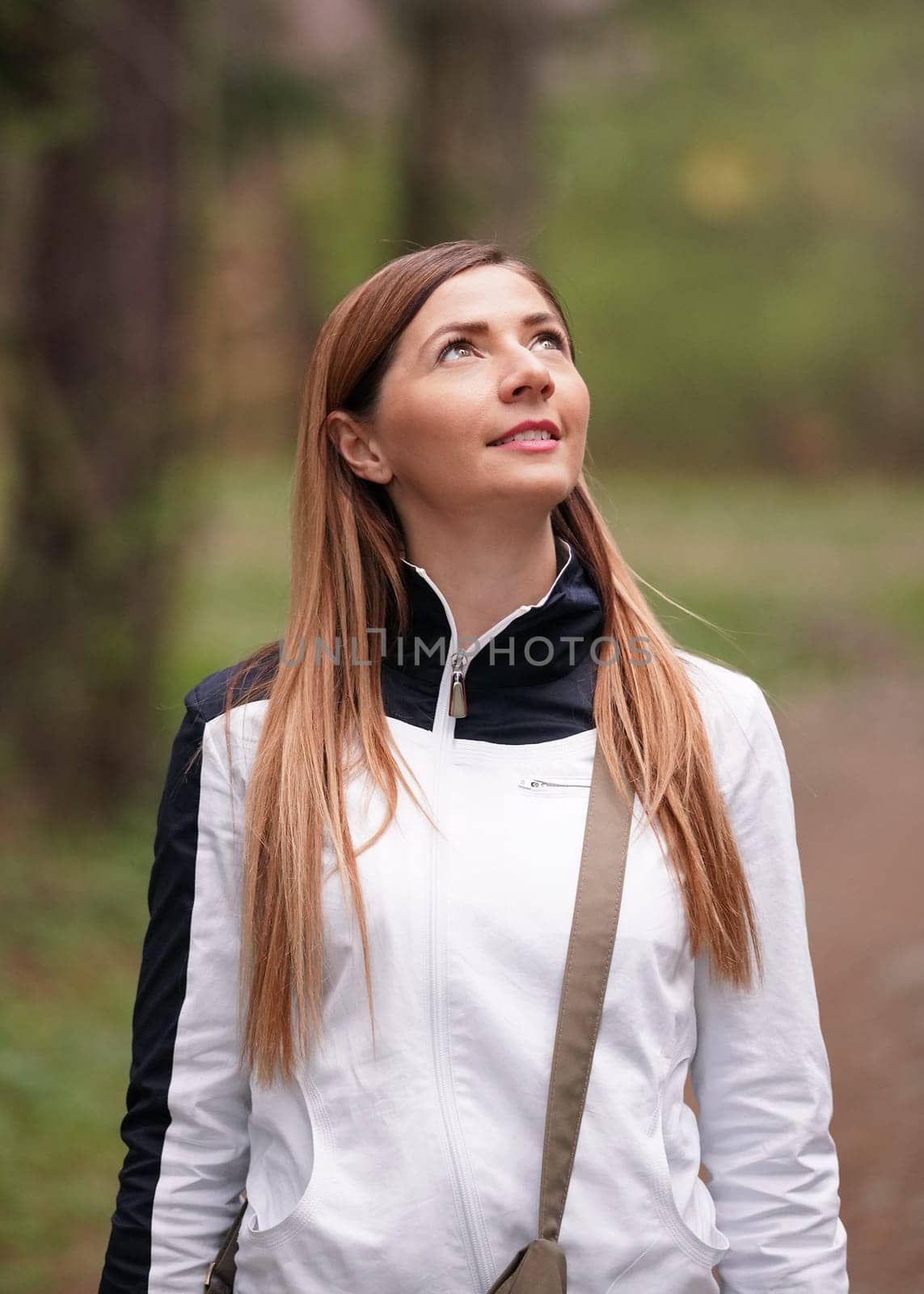 Portrait of young woman wearing white sport jacket, looking up, blurred trees in background by Ivanko