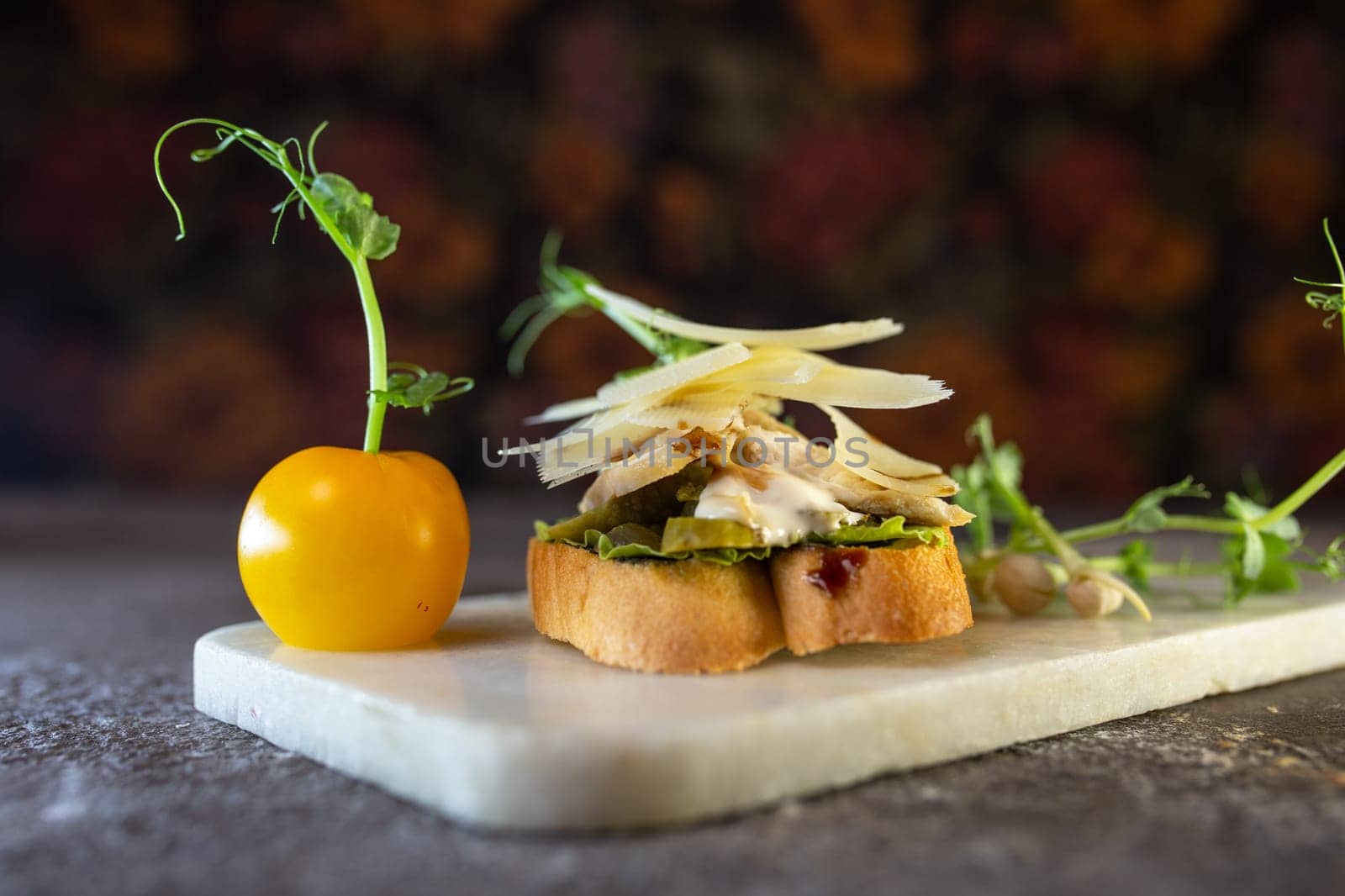 A bruschetta with chicken, pesto, and parmesan on a white marble board. The bruschetta is garnished with microgreens and a pea pod.