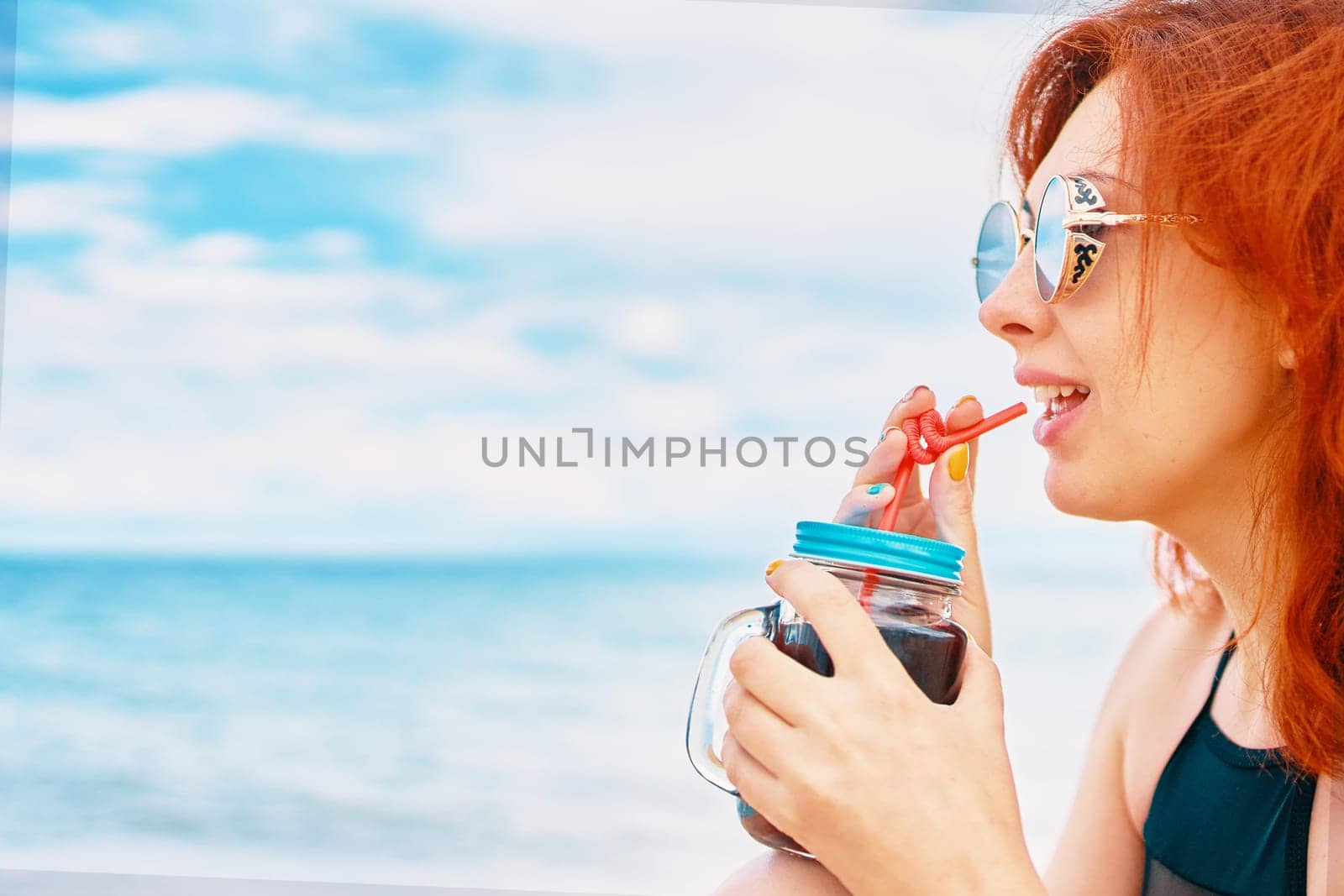 Red-haired woman in a swimsuit is sitting on beach with a cocktail and look away. Tropical alcohol beverage. Summer day with cloudy sky. Calm lady with sunglasses on vacation. Holidays at the sea.
