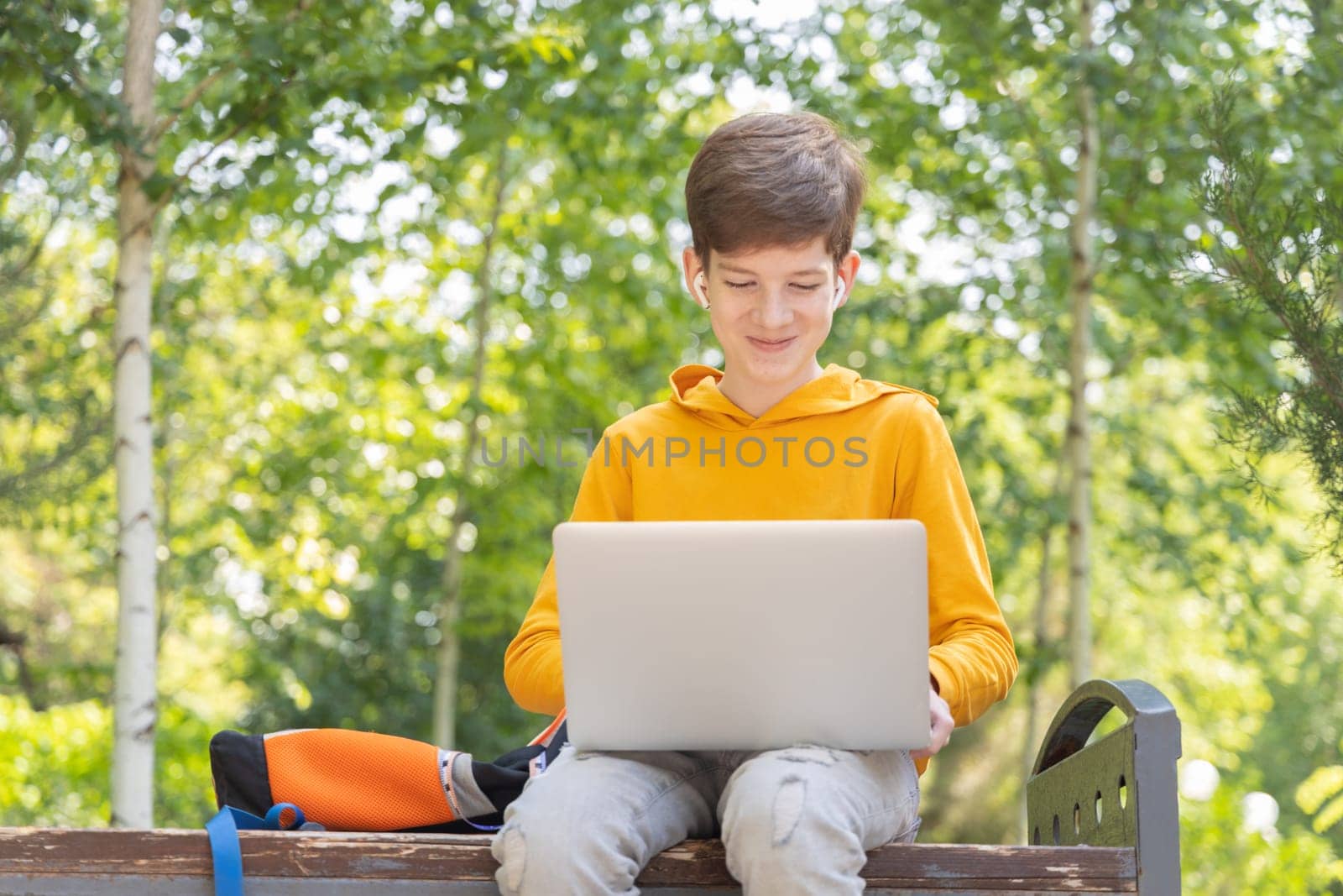 Smiling teenager boy working on laptop. Holding and using a laptop for networking on a sunny spring day, outdoors. by Ri6ka