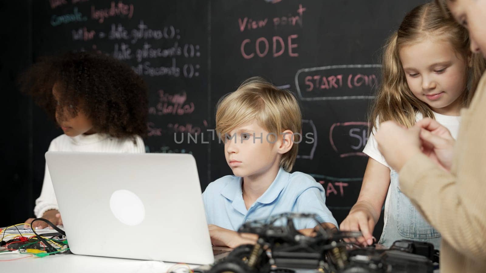 Closeup of boy using laptop programing engineering code and writing program while group of smart diverse student standing surrounded by friend in STEM technology classroom at blackboard. Erudition.