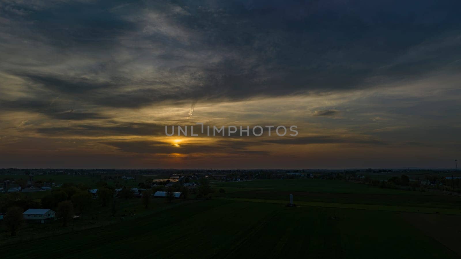 Dramatic Sunset Casting A Warm Glow Over A Rural Landscape With Silhouetted Trees And Buildings Under A Cloud-Streaked Sky.