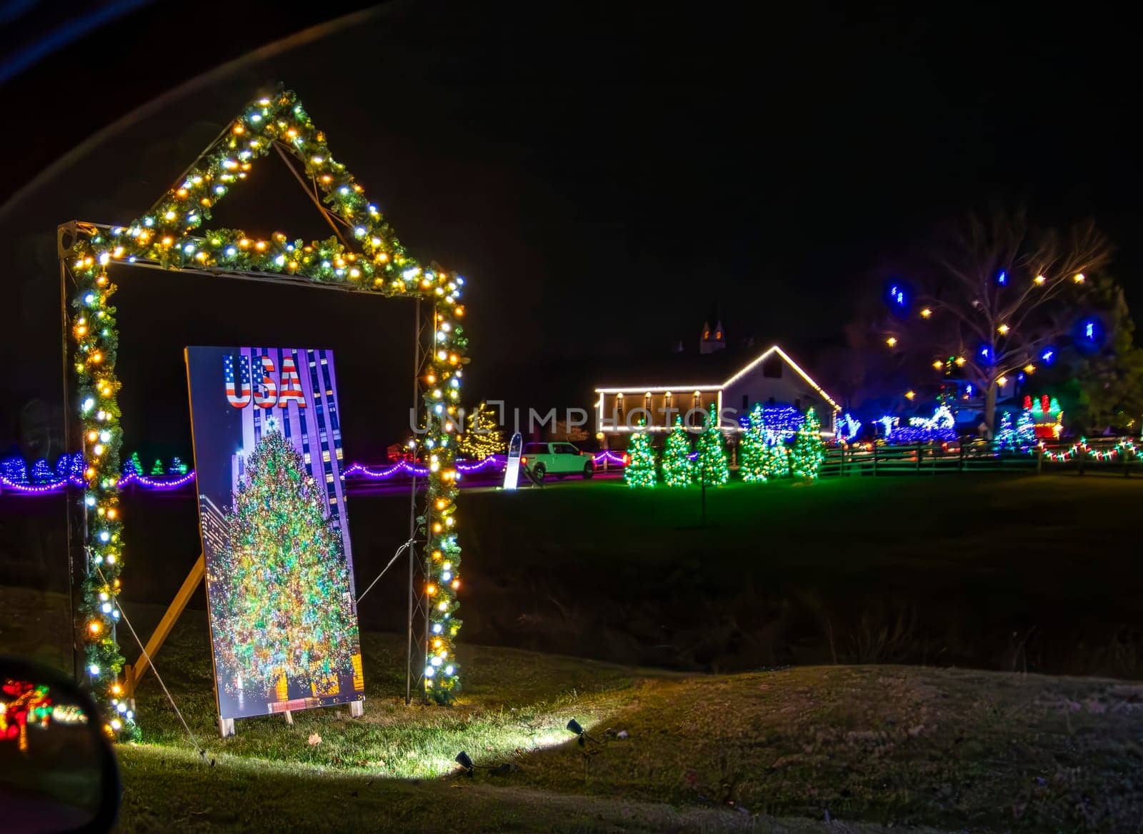 Night View Of A Colorful Holiday Display Featuring A Lit Archway, A Sign With A Christmas Tree Image, And A Streetlamp Surrounded By Glowing Red Bushes.