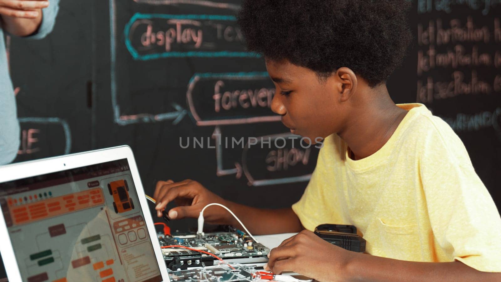 African boy learning to use electronic tool while laptop display engineering code or prompt while sitting in front of blackboard with web develop plan written at STEM technology class. Edification.
