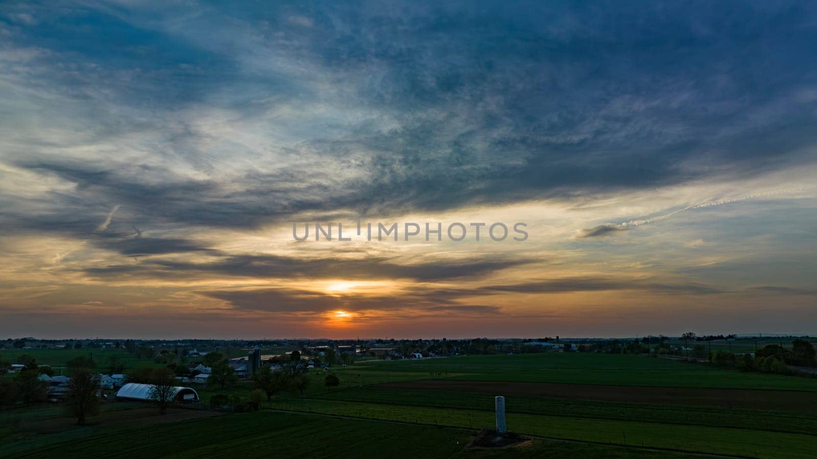 Aerial View of Dramatic Sunset Casting A Warm Glow Over A Rural Landscape With Silhouetted Trees And Buildings Under A Cloud-Streaked Sky.