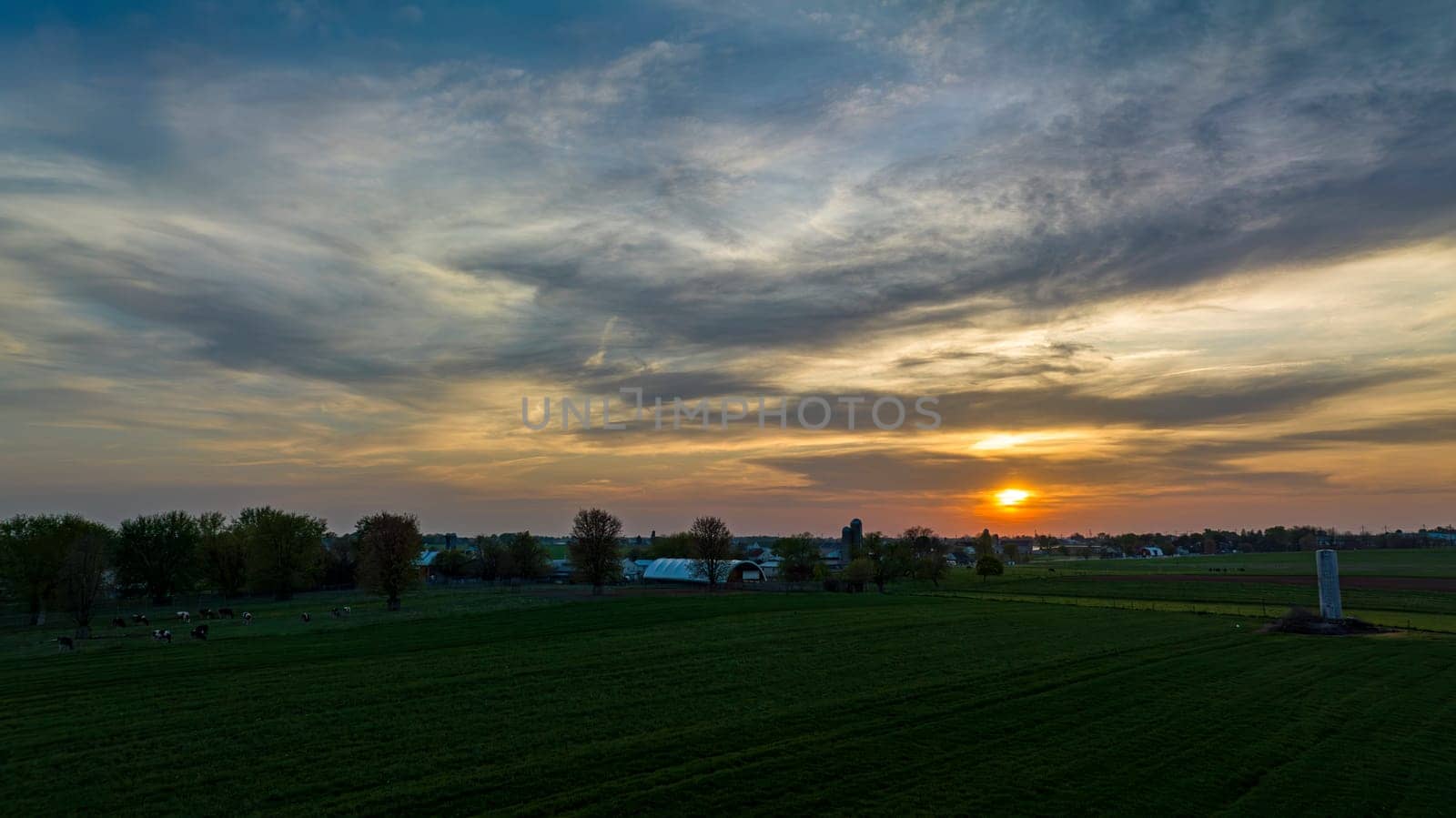 Aerial View of Dramatic Sunset Casting A Warm Glow Over A Rural Landscape With Silhouetted Trees And Buildings Under A Cloud-Streaked Sky.