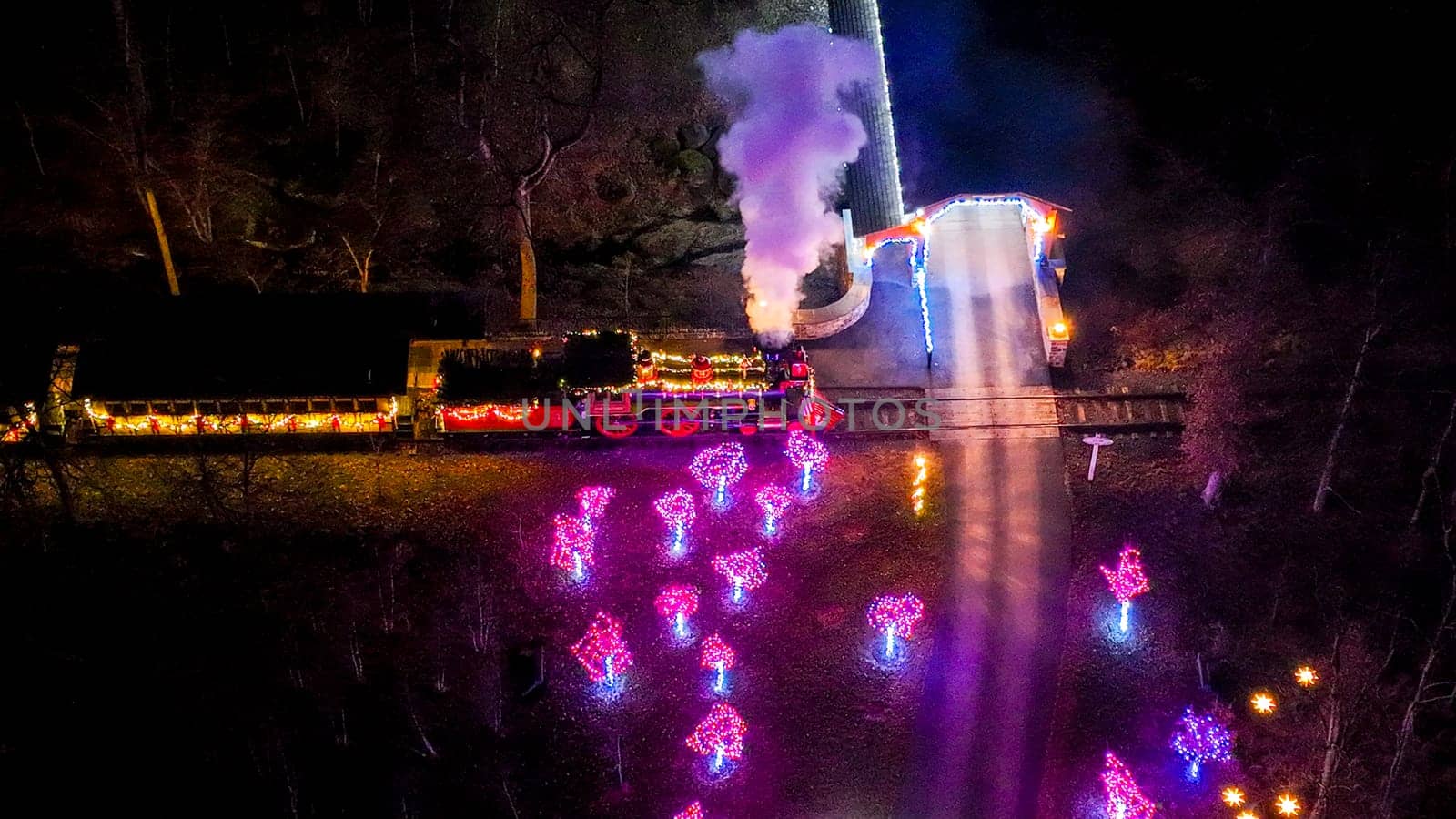 Direct Overhead View Of A Vintage Train Decorated With Christmas Lights Emitting Steam At Night, Surrounded By Trees With Glowing Lights.