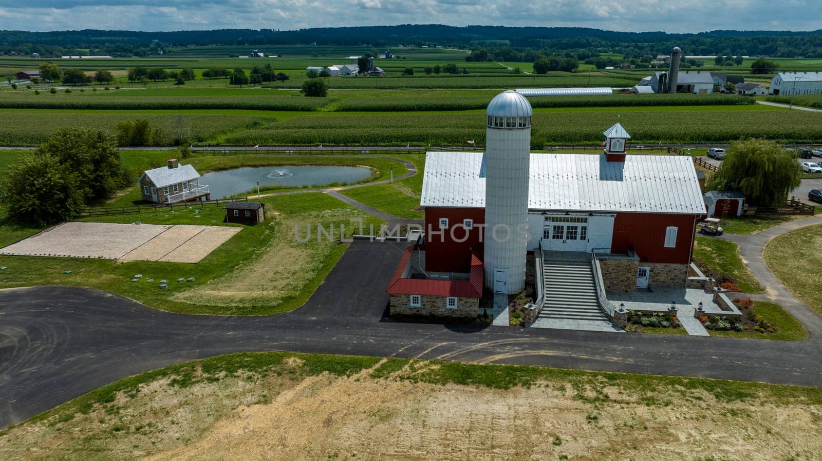 Overlooking A Farmstead Featuring A Majestic Red Barn With Silo, An Adjacent Pond With Geese, Winding Asphalt Paths, And Lush Green Crop Fields Under A Blue Sky With White Clouds.