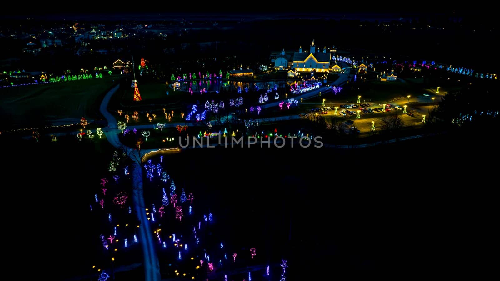 High-Angle Night View Of A Lively Holiday Lighting Display With A Road Leading To A Central Pond And Clustered Lit Structures.