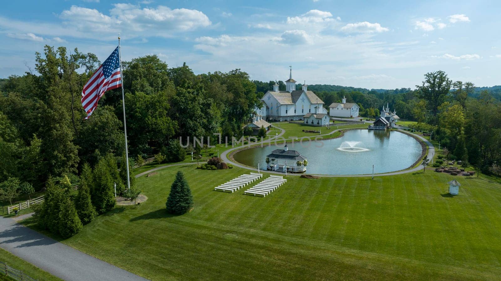 Aerial View Of A Grand White Building With A Grey Roof Surrounded By Lush Green Lawns, A Large Reflective Pond With Fountains, And A Majestic Flagpole Bearing The American Flag.