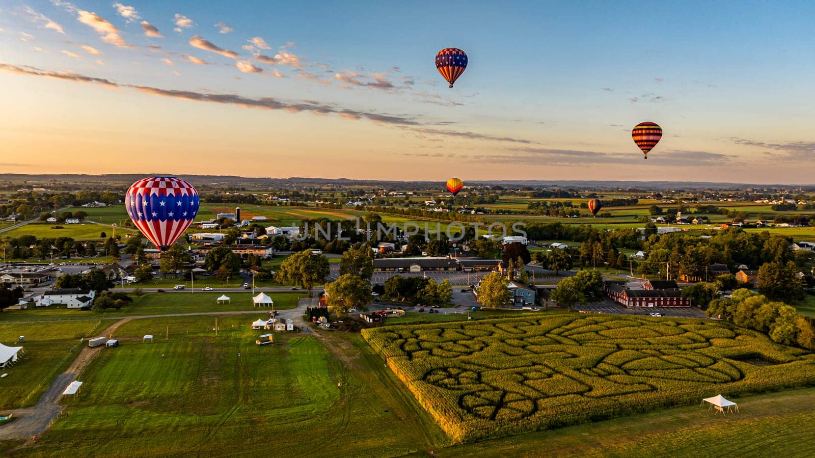An Aerial View of Multiple Hot Air Balloons Soar Over A Picturesque Landscape Featuring A Corn Maze, Town, And Fields During A Golden Hour Sunset.