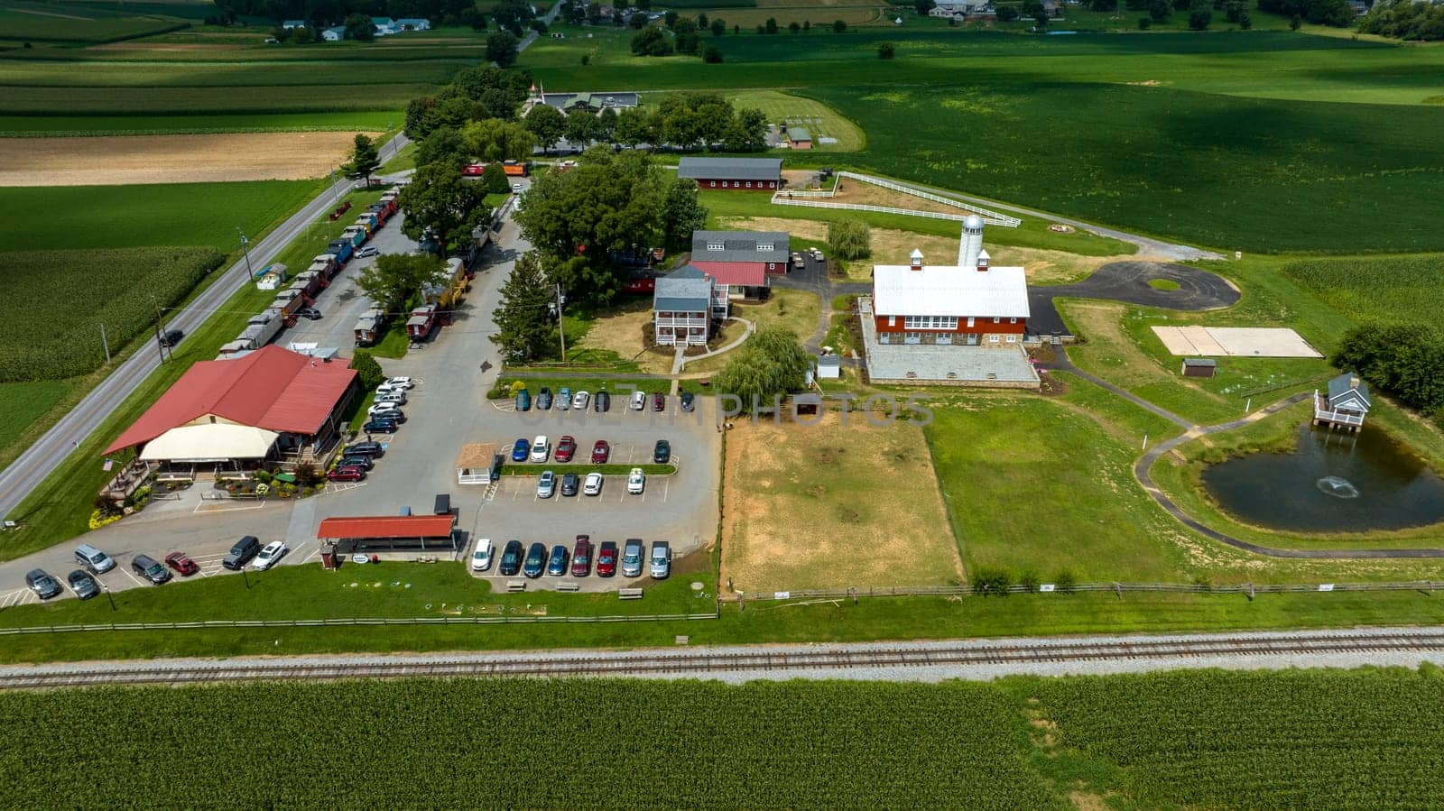 Overhead View Of A Bustling Country Market With A Red Roof, Adjacent To A Farm, Parking Lot Full Of Cars, Railroad Tracks, And Surrounding Green Fields.