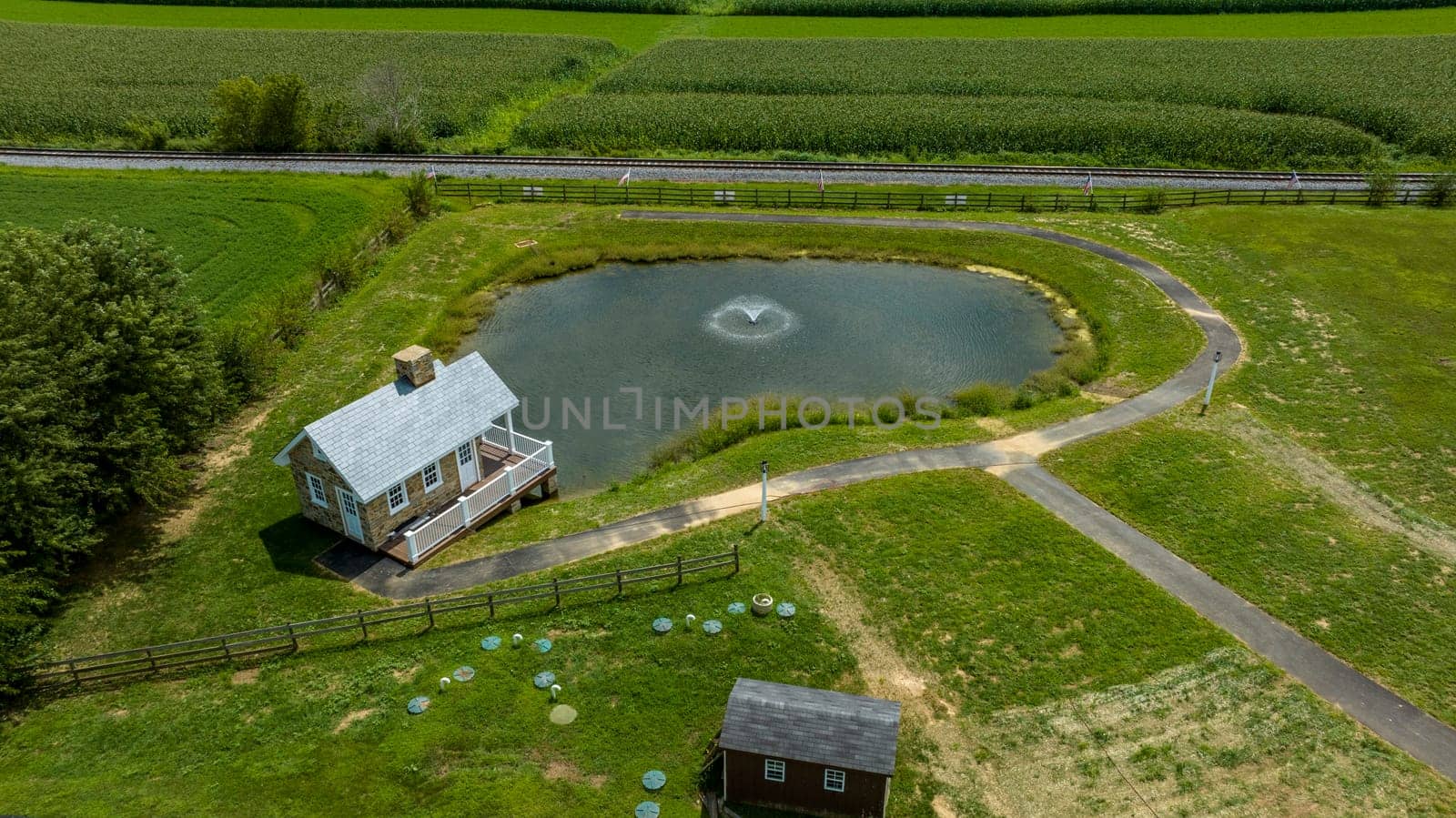 Aerial View Of A Small Pond With A Fountain Next To A Two-Story House And A Curving Path With Lush Farmland And A Railroad Track In The Background.