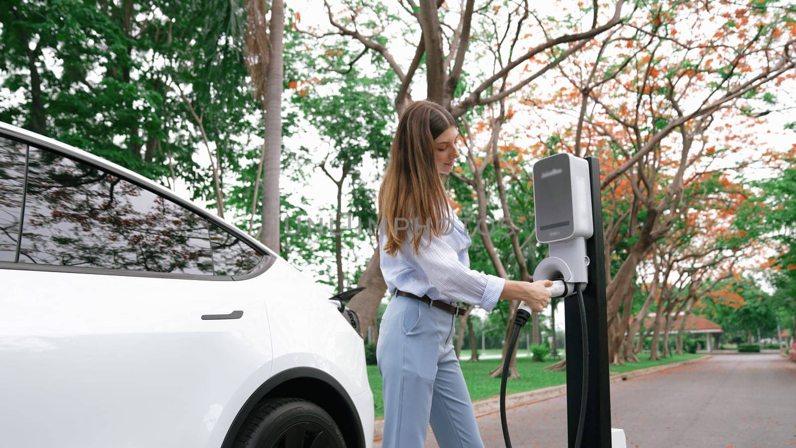 Woman recharging battery for electric car during road trip travel EV car in autumanl season natural forest or national park. Eco friendly travel during vacation and holiday. Exalt