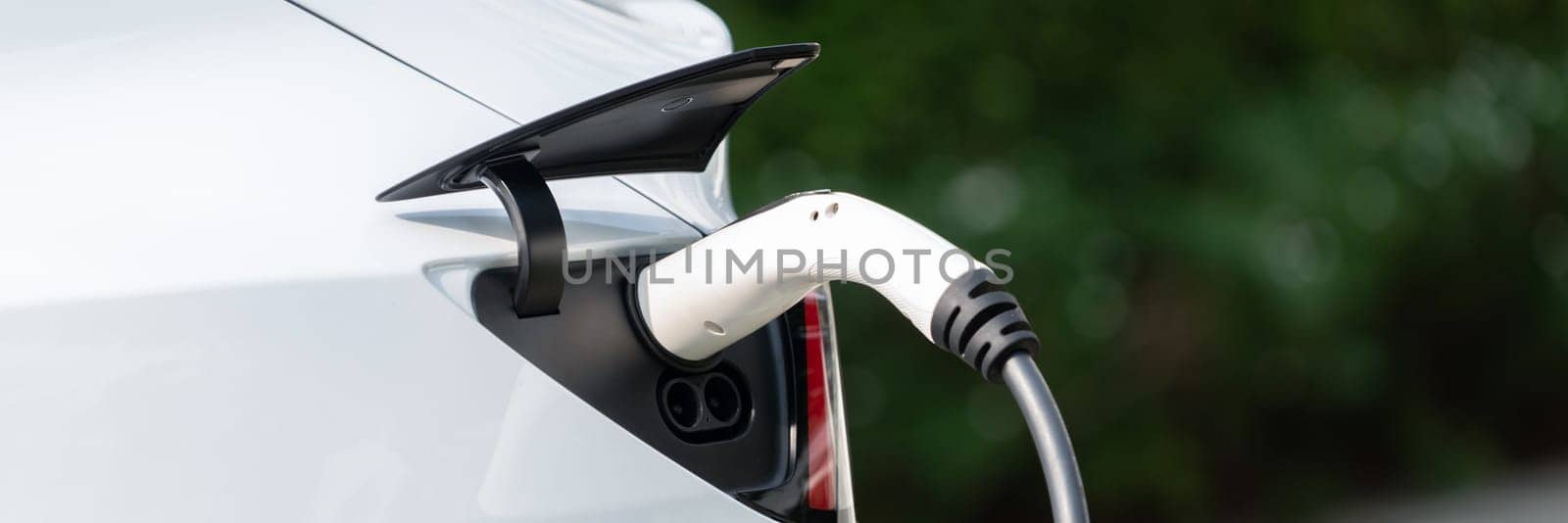 Closeup EV charger plug handle attached to electric vehicle port, recharging battery from charging station. Modern designed EV car and clean energy sustainability for better future.Panorama Synchronos