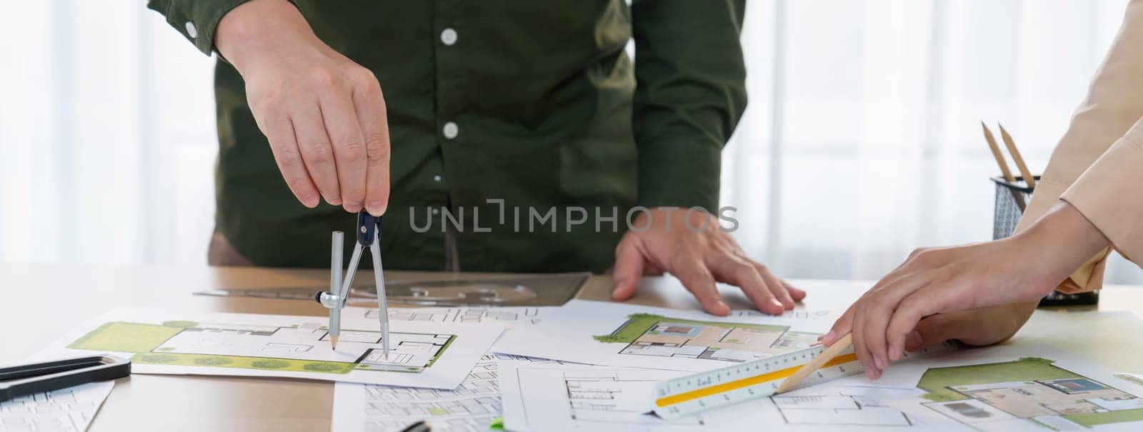 A cropped image of professional engineer using divider to measure blueprint at meeting table with blueprint, laptop and architectural equipment scatter around. Closeup. Delineation.