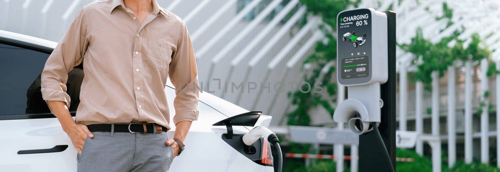 Young man recharge electric car's battery from charging station in city commercial parking lot. Rechargeable EV car for sustainable environmental friendly urban travel. Panorama Expedient