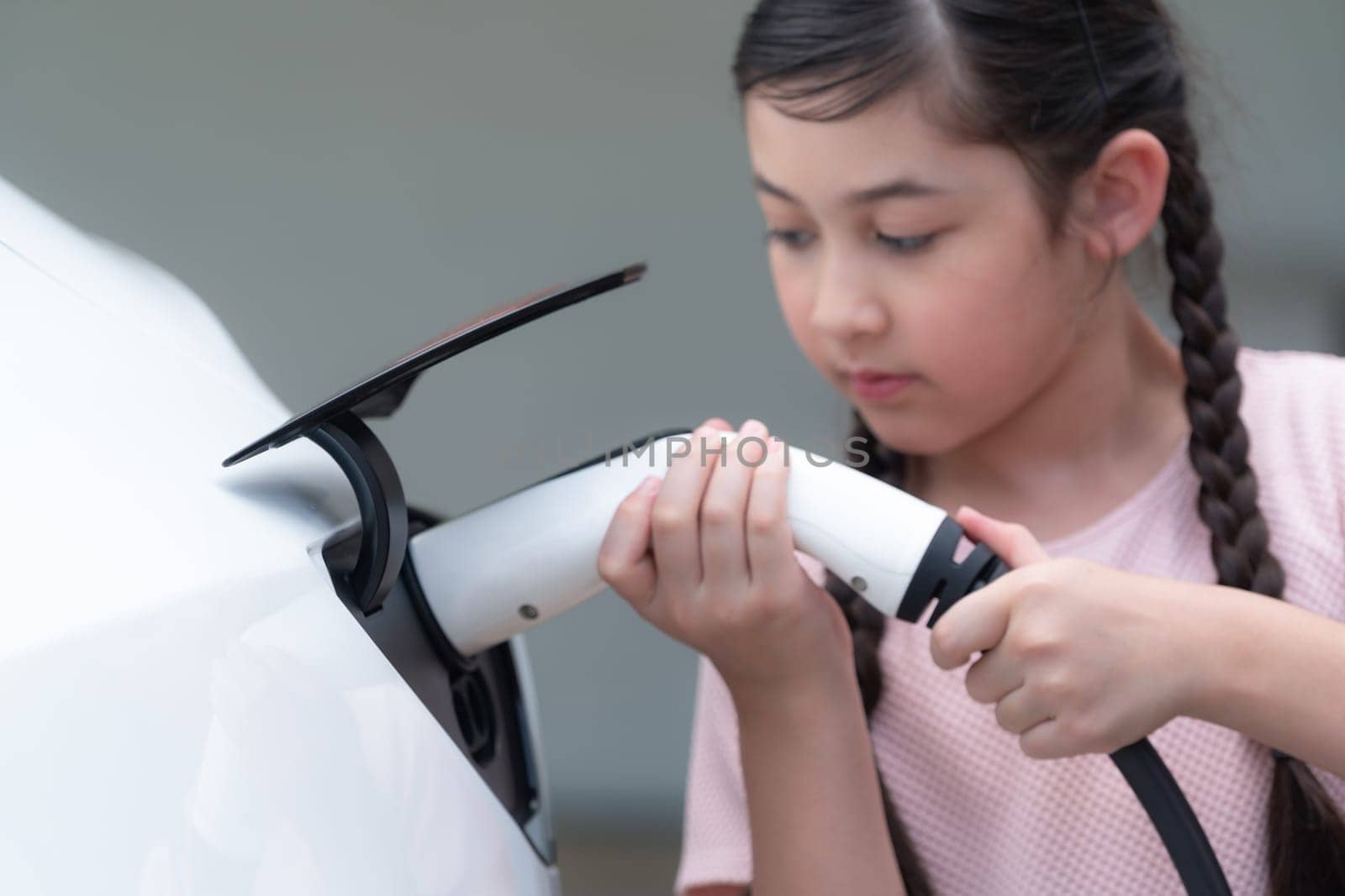 Happy little young girl learn about eco-friendly and energy sustainability as she recharge electric vehicle from home EV charging station. EV car and sustainable future generation concept. Synchronos