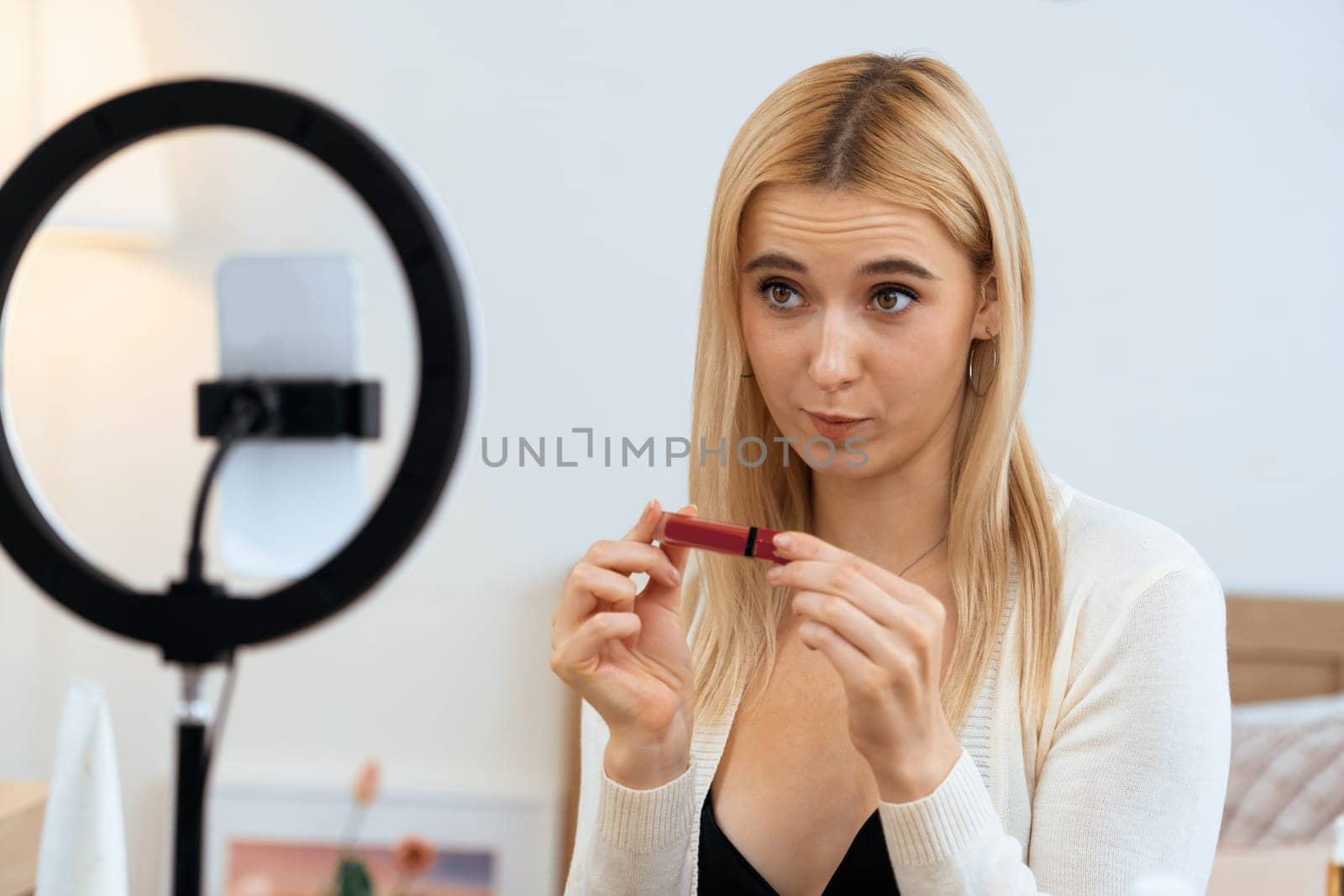 Young woman making beauty and cosmetic tutorial video content for social media. Beauty blogger using camera and light ring while showing how to apply liquid lipstick to audience or follower. Blithe