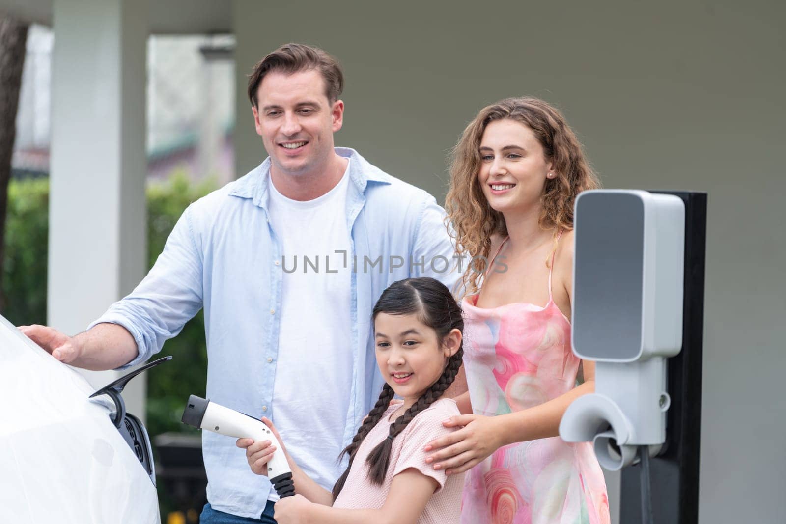 Happy little young girl learn about eco-friendly and energy sustainability as she help her family recharge electric vehicle from home EV charging station. EV car and modern family concept. Synchronos