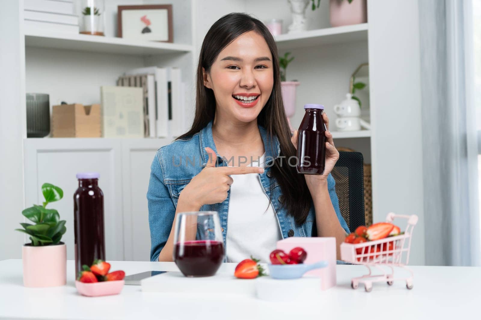 Smiling young beautiful Asian reviewing tasty mixed strawberry and cherry juice detox drinks, showing healthy product to promote for special promotion selling on social media online record. Stratagem.