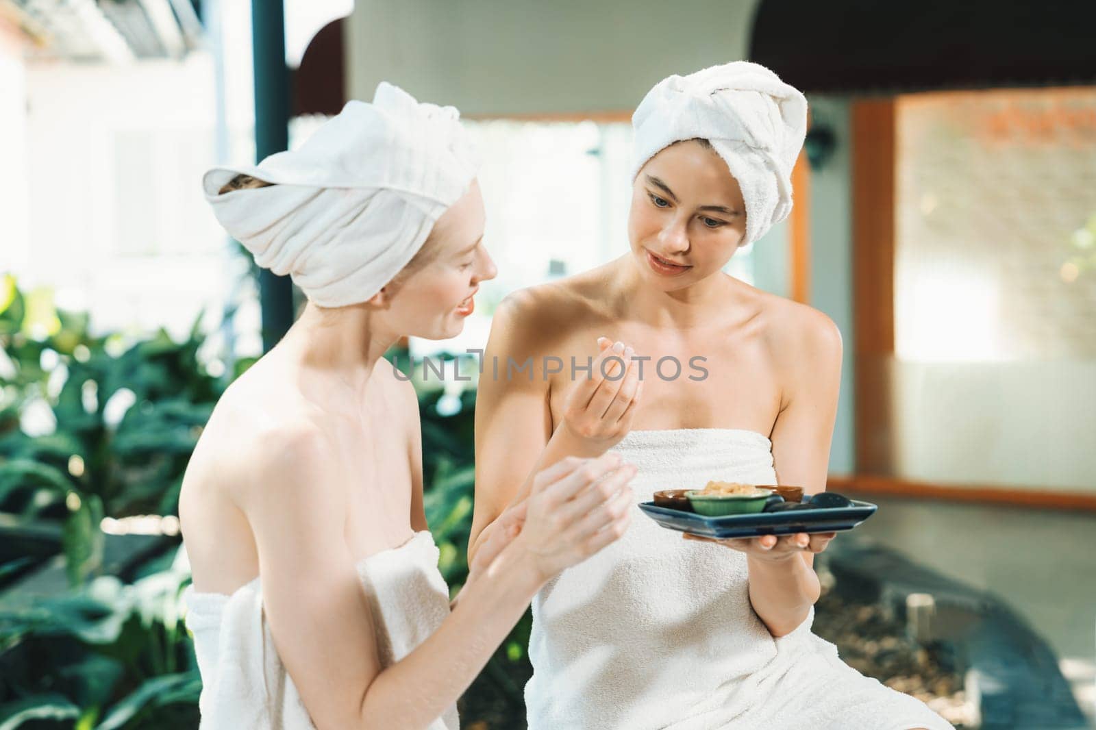 Portrait of two attractive woman in towel giggling during hold the herbal bowl surrounded by natural spa environment. Pretty girls with beautiful skin using herbal scrub at spa salon.Tranquility.
