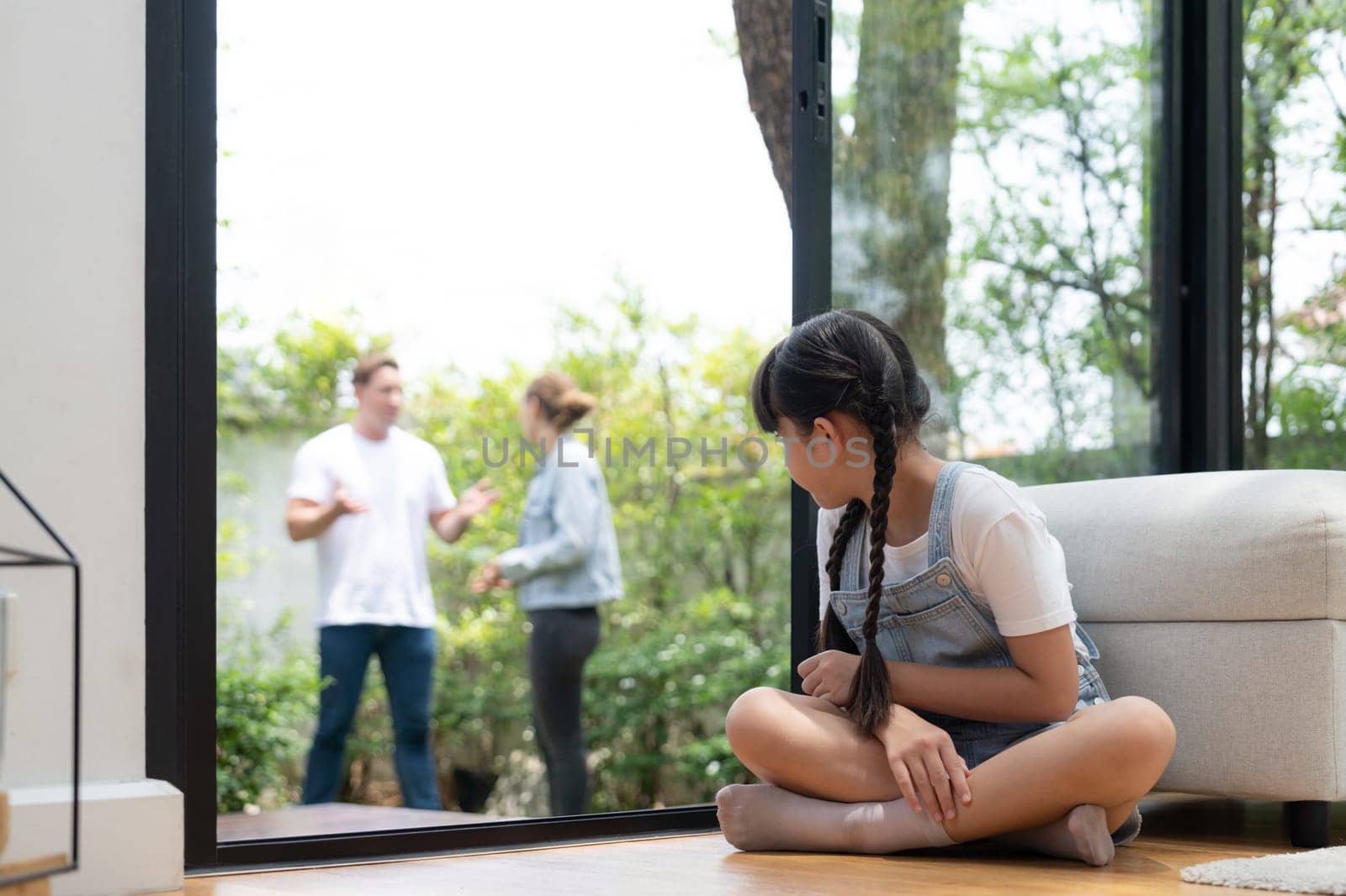 Stressed and unhappy young girl huddle in corner crying and sad while her parent arguing in background. Domestic violence at home and traumatic childhood develop to depression. Synchronos