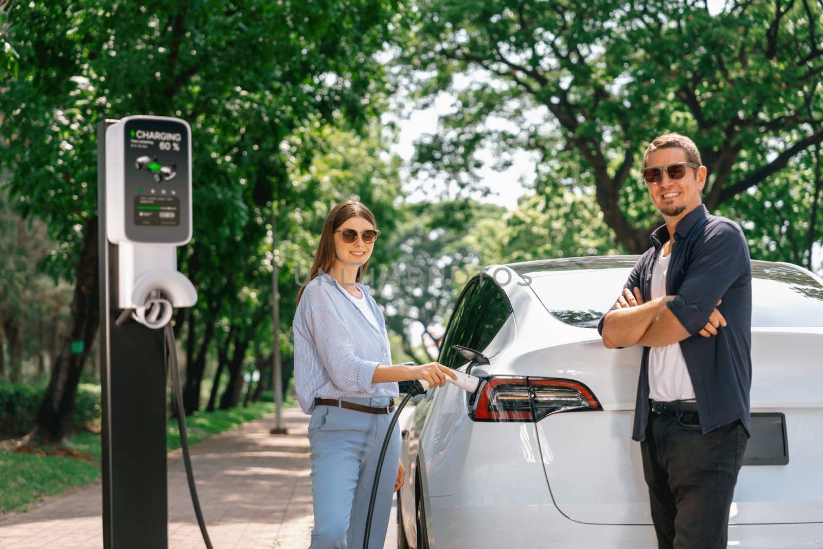 Lovely young couple wearing sun glasses recharging battery for electric car during road trip travel EV car in natural forest or national park. Eco friendly travel during vacation and holiday. Exalt