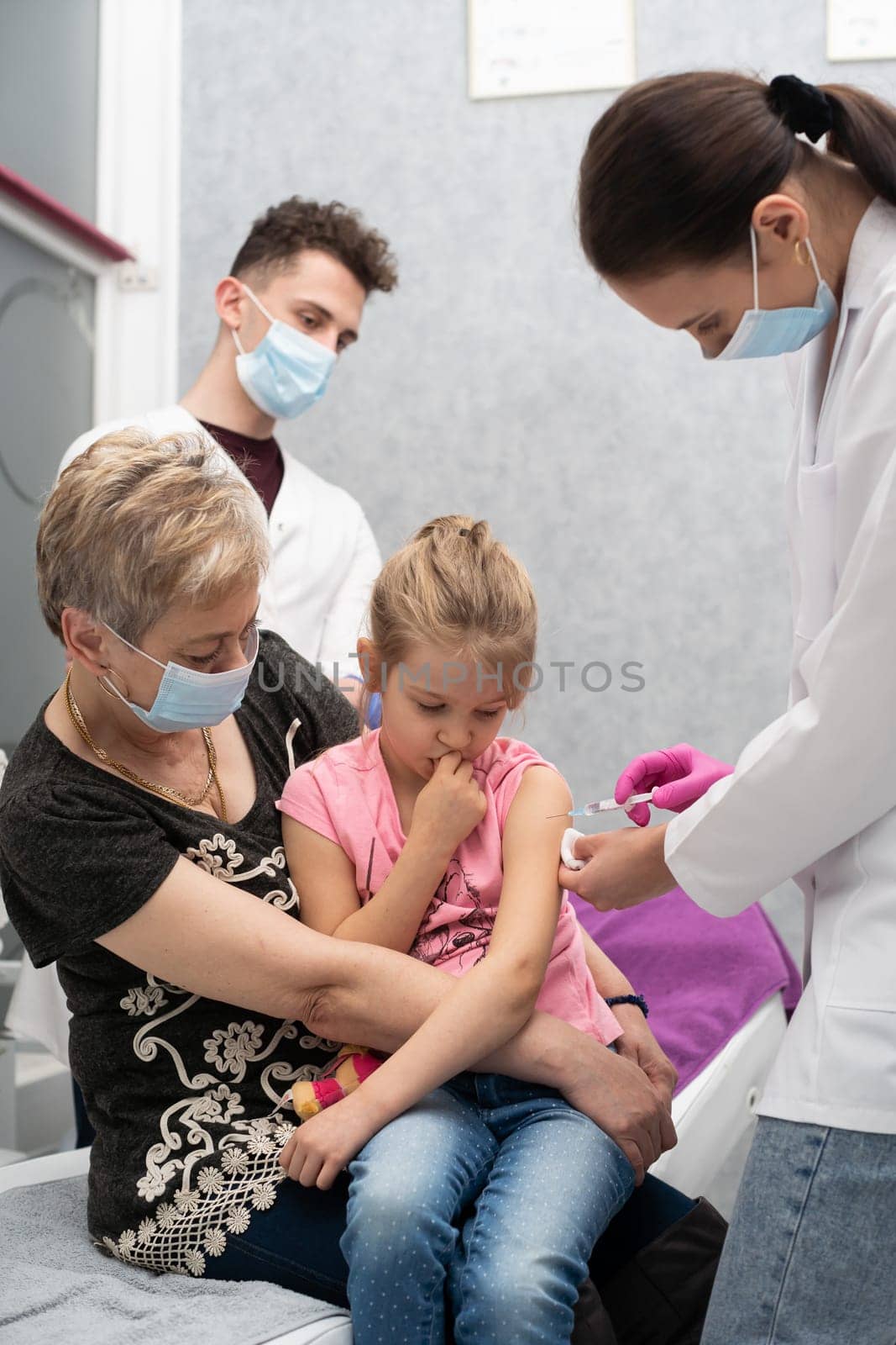 Grandma took her granddaughter on her lap so that the child would not be afraid of the injection made by the young nurse. A young nurse is injecting a new vaccine. Safe vaccination of children.