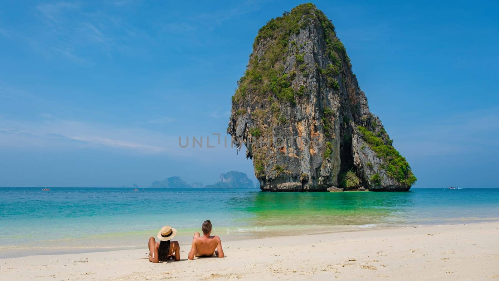 Two individuals are relaxing on a sandy beach with a majestic rock formation in the background, overlooking the vast ocean with clear blue skies and a few clouds above Couple vacation Krabi Thailand