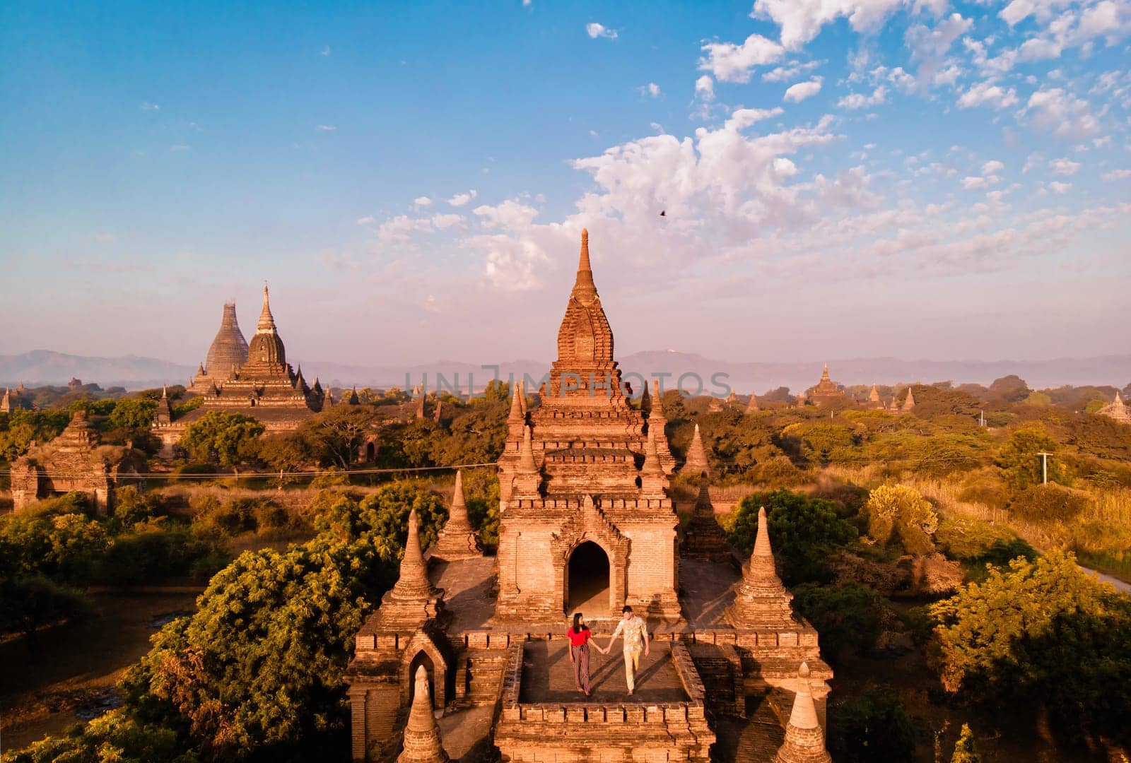An aerial view of a temple set amidst lush trees with a backdrop of a clear blue sky and fluffy cumulus clouds, creating a tranquil natural landscape, couple on vacation in Myanmar