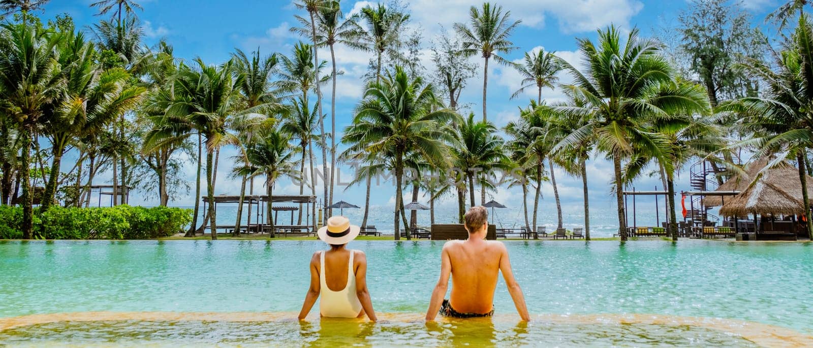 A couple relaxing in a pool, palm trees all around them under the blue sky by fokkebok