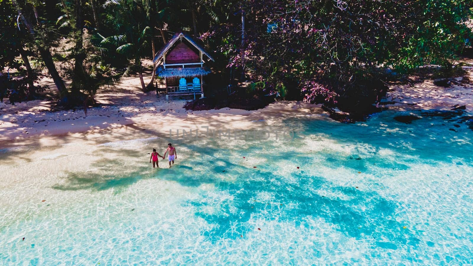 top view at a couple on a tropical beach with palm trees and a wooden bamboo hut bungalow. Koh Wai Island Trat Thailand. drone view at young man and woman on the beach