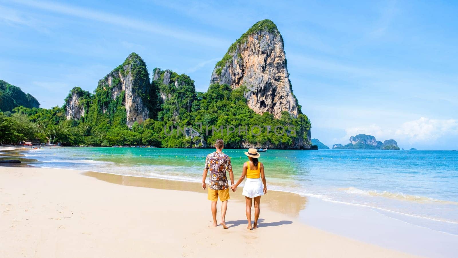 a diverse couple of men and women on the beach, an Asian woman and a caucasian man walking on the beach of Railay Beach in Thailand with limestone cliffs in the background