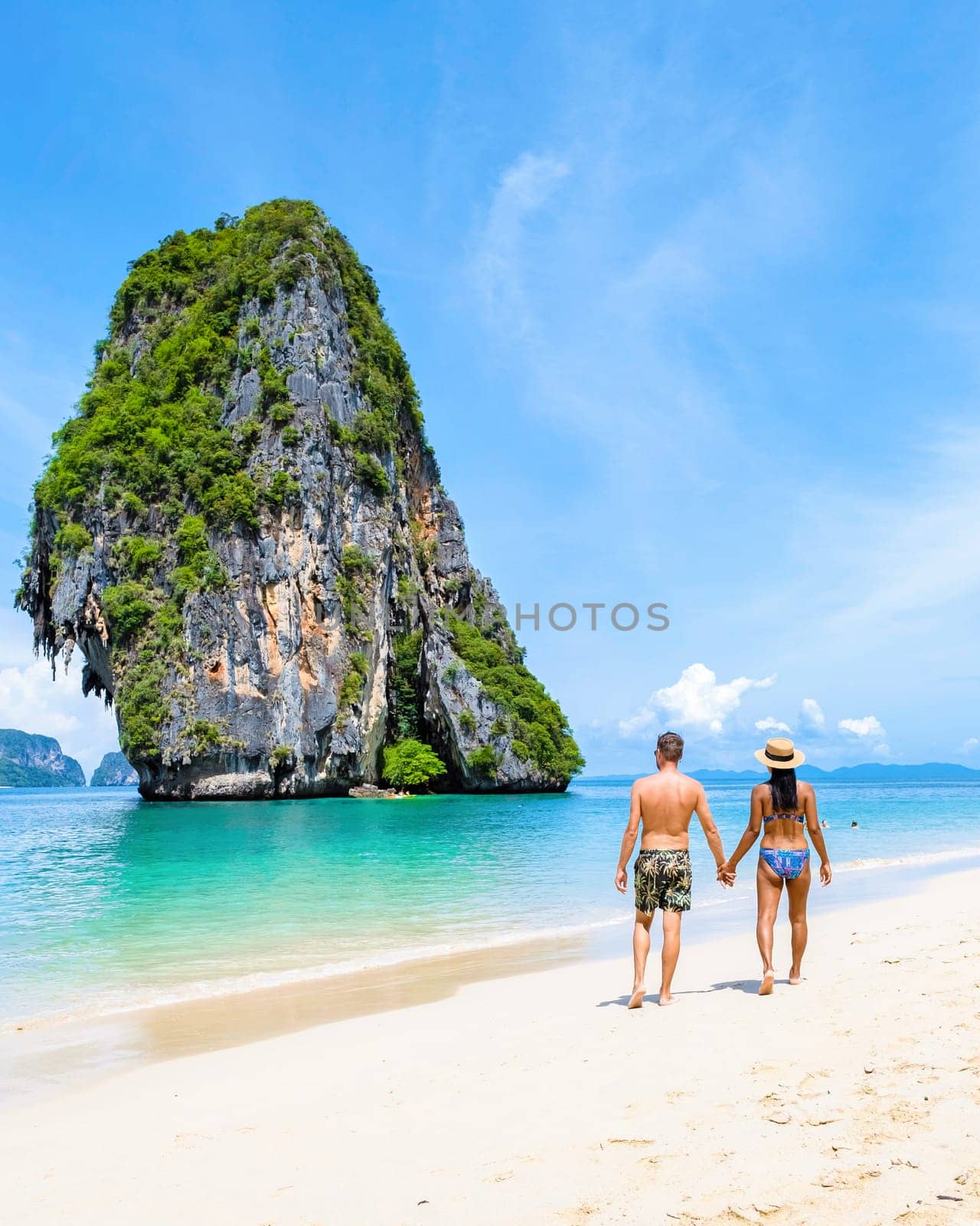 diverse couple of men and women on the beach of Railay Krabi Thailand, an Asian woman in a bikini and a Caucasian man in swim short walking at the beach with a huge limestone cliff in the background
