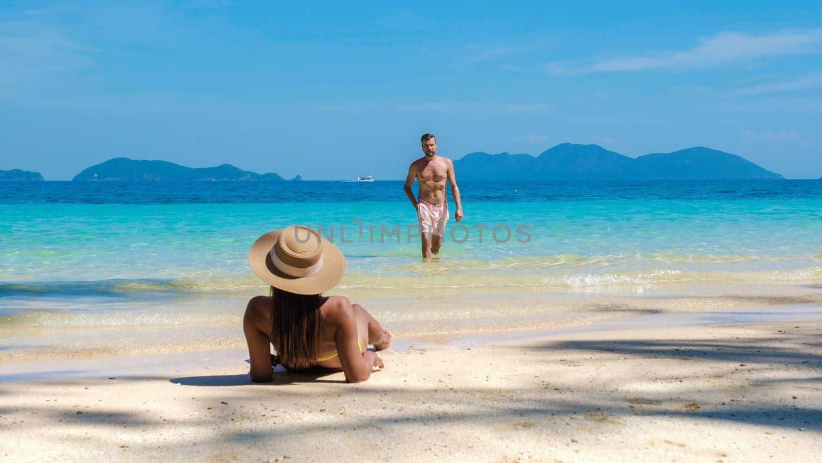 a young couple of men and women on a tropical beach during a luxury vacation in Thailand, an Asian woman with a summer hat lying down on the beach while caucasian men walking in the ocean