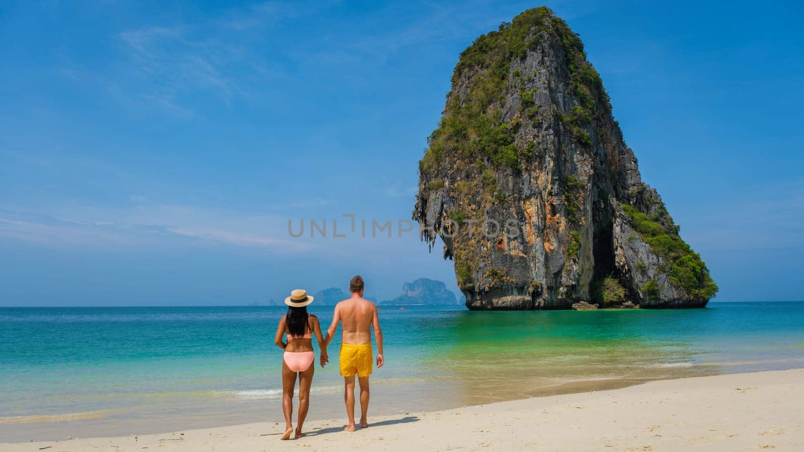 couple of men and women on the beach, an Asian woman and a caucasian man walking on the beach of Railay Beach in Thailand with limestone cliffs in the background, woman in bikini man in swim short