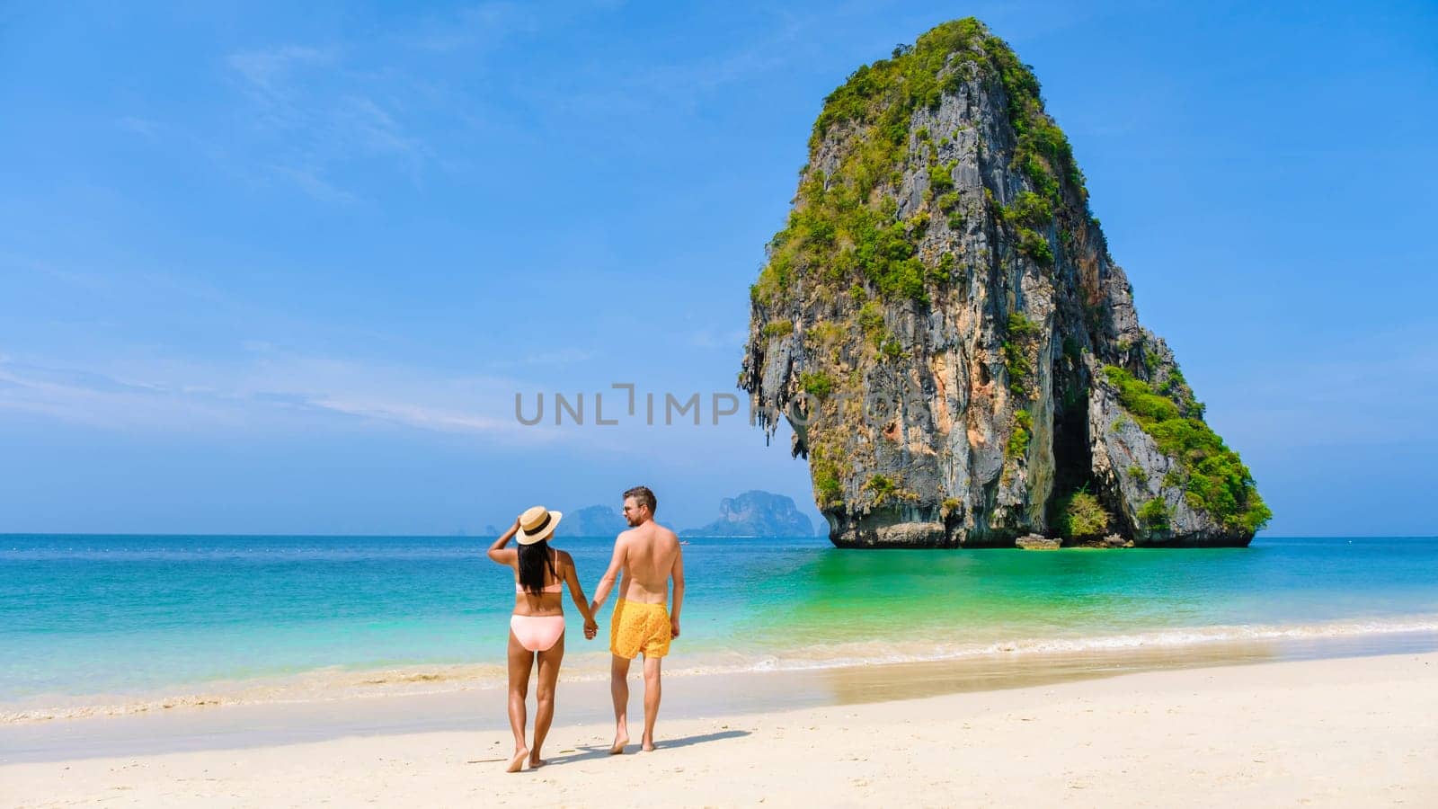 a diverse couple of men and women on the beach, an Asian woman in bikini and a caucasian man in swim short walking on the beach of Railay Beach in Thailand with limestone cliffs in the background