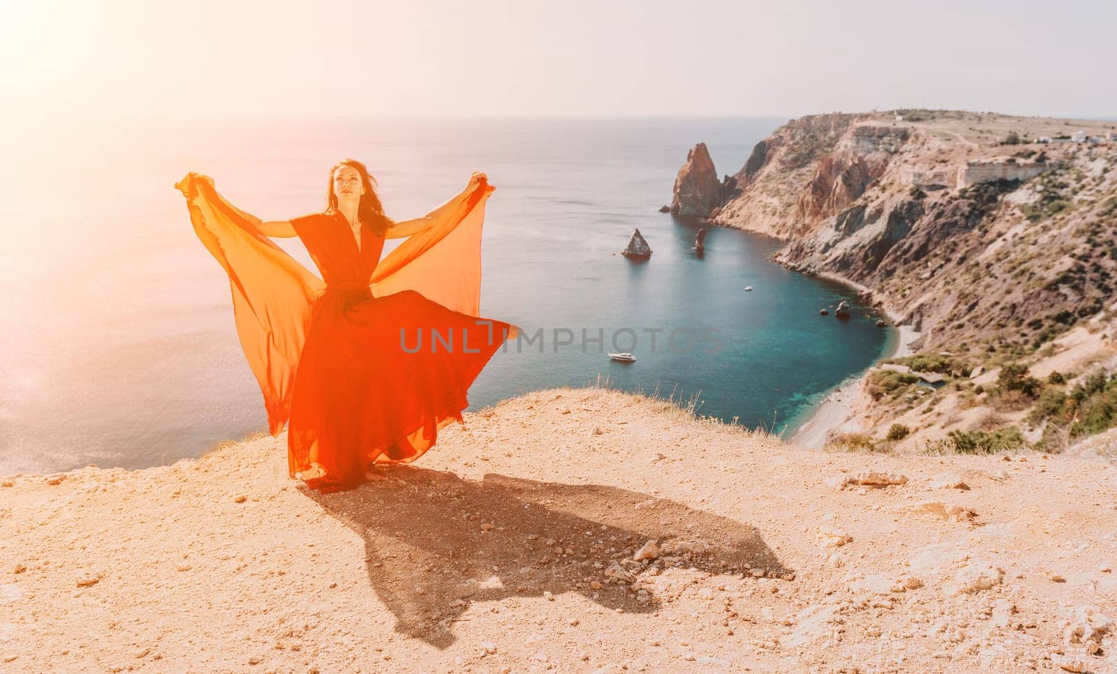 Side view a Young beautiful sensual woman in a red long dress posing on a rock high above the sea during sunrise. Girl on the nature on blue sky background. Fashion photo.